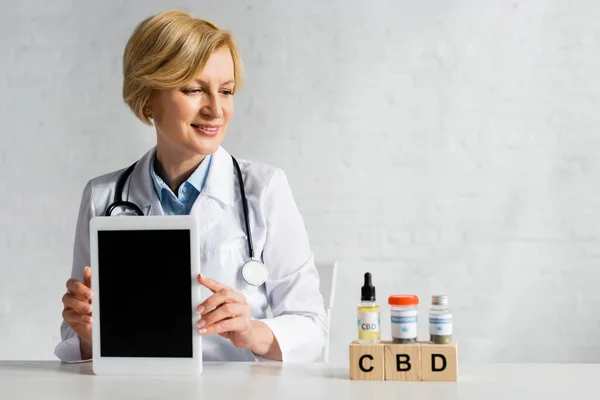 Happy doctor holding digital tablet with blank screen near cubes with cbd lettering and drugs — Stock Photo