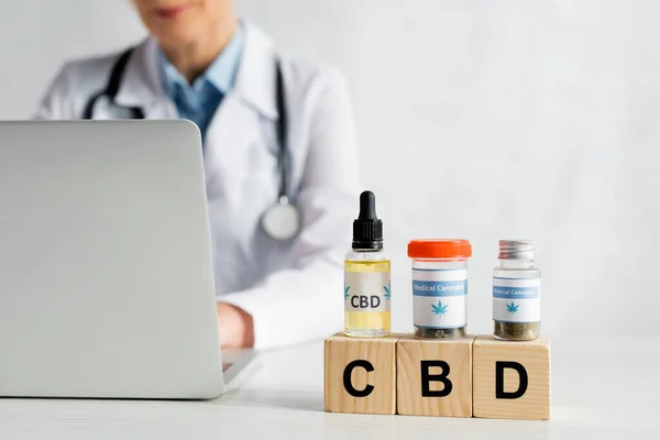 Vista ritagliata del medico utilizzando laptop vicino a cubi con scritte cbd e bottiglie con cannabis medica — Foto stock