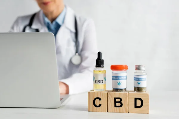 Обрезанный вид врача с помощью ноутбука рядом с кубиками с надписью CBD и бутылки с медицинским каннабиса — стоковое фото