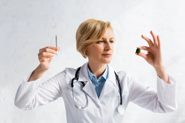 Mature doctor looking at dried marijuana while holding rolled joint in clinic — Stock Photo