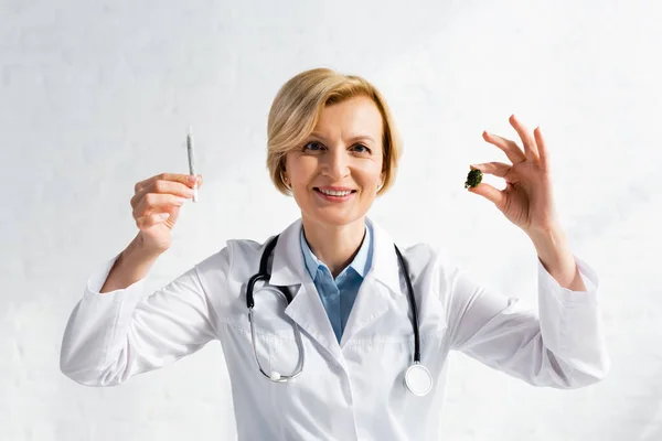 Happy and mature doctor holding dried marijuana and rolled joint in clinic — Stock Photo