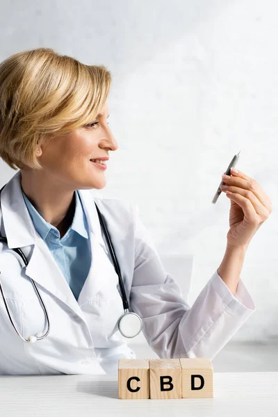 Happy and mature doctor holding rolled joint near cubes with cbd lettering — Stock Photo