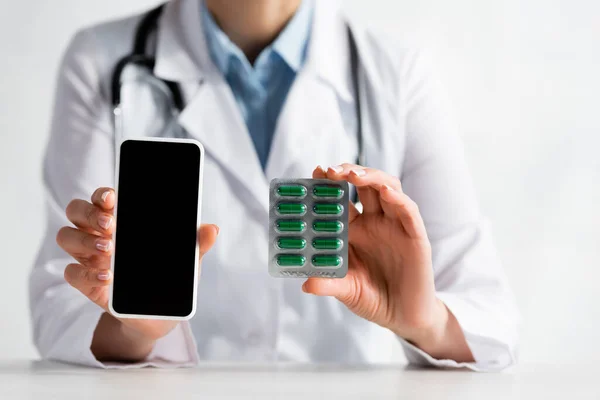 Vista recortada del médico maduro sosteniendo teléfono inteligente con pantalla en blanco y blister con pastillas - foto de stock