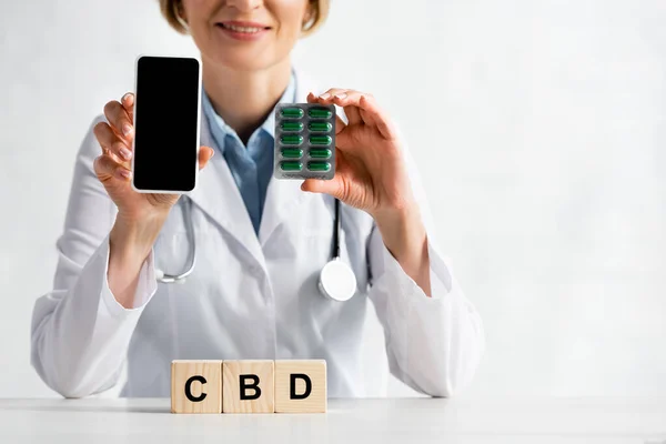 Vista recortada de feliz médico sosteniendo teléfono inteligente con pantalla en blanco y blister paquete con pastillas cerca de cubos con letras cbd - foto de stock