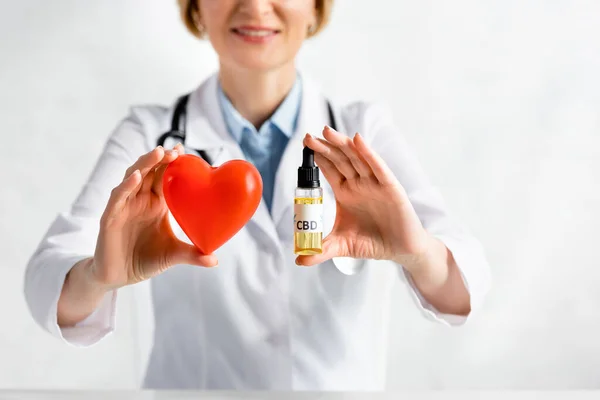 Обрезанный вид веселого и зрелого врача, держащего бутылку с надписью CBD и красным сердцем в клинике — стоковое фото