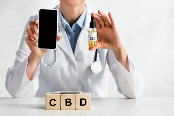 Обрезанный вид врача, держащего смартфон с пустым экраном и бутылкой масла возле кубиков с надписью cbd — стоковое фото