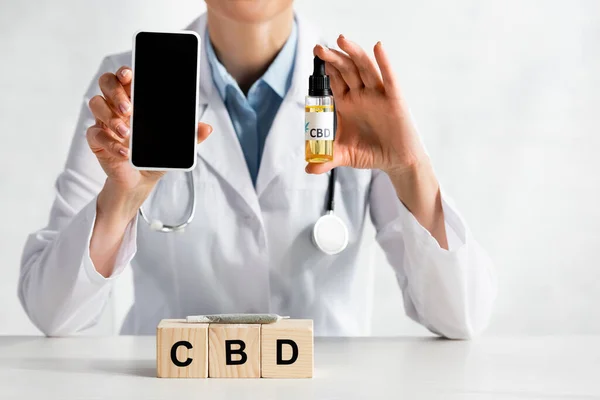 Vue recadrée du médecin tenant smartphone avec écran vierge et bouteille d'huile près de cubes avec lettrage cbd — Photo de stock