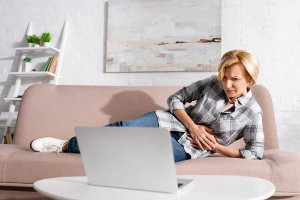Mature woman suffering from stomach ache and lying on sofa near laptop — Stock Photo
