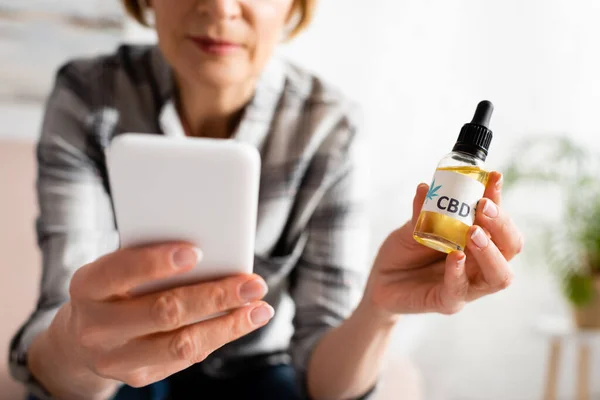 Cropped view of mature woman holding bottle with cbd lettering and using smartphone — Stock Photo