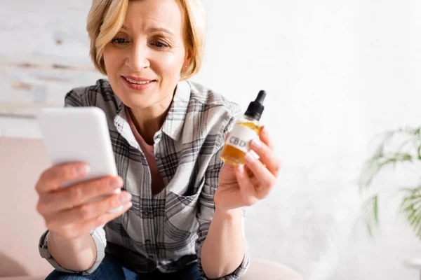 Selektiver Fokus einer glücklichen und reifen Frau, die eine Flasche mit cbd-Schriftzug in der Hand hält und das Smartphone benutzt — Stockfoto