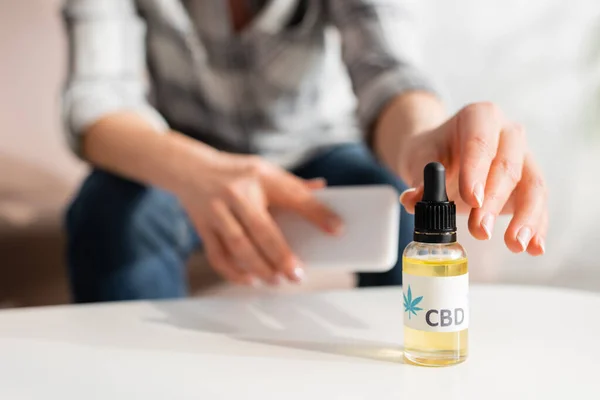 Abgeschnittene Ansicht einer reifen Frau, die eine Flasche mit cbd-Schriftzug nimmt und ihr Smartphone zu Hause hält — Stockfoto