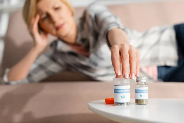 Selective focus of mature woman touching bottle with medical cannabis lettering while lying on sofa — Stock Photo