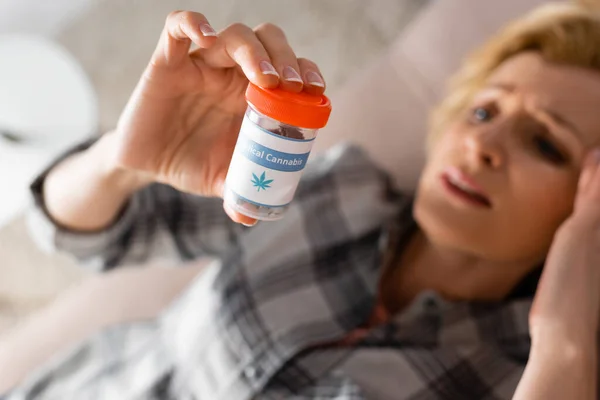 Selective focus of mature woman holding bottle with medical cannabis lettering and with having headache — Stock Photo