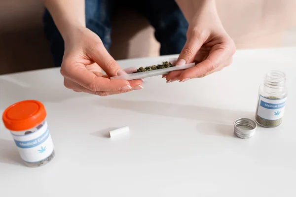 Vista recortada de una mujer madura sosteniendo papel con hierba seca cerca de botellas con letras de cannabis medicinal - foto de stock