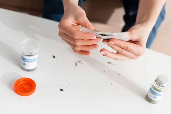 Vista recortada de una mujer madura sosteniendo papel con marihuana seca cerca de botellas con letras de cannabis medicinal - foto de stock