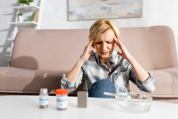 Reife Frau mit Migräne in der Nähe von Flaschen mit medizinischem Cannabis und Aschenbecher — Stockfoto