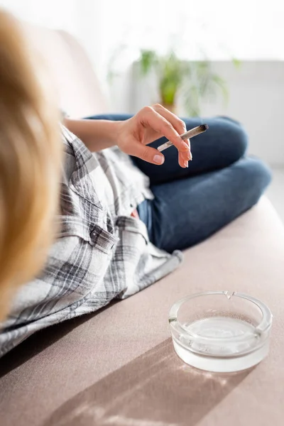 Selective focus of mature woman lying on sofa and holding joint with legal marijuana near ashtray — Stock Photo