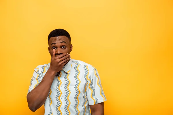 Hombre afroamericano asustado cubriendo la boca con la mano sobre fondo amarillo - foto de stock