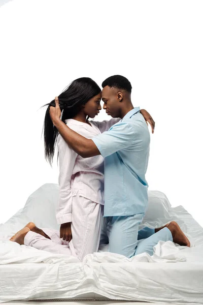 Vista lateral de pareja afroamericana abrazándose en la cama en pijama aislado en blanco - foto de stock
