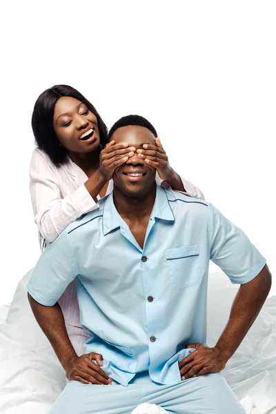 Happy african american woman covering eyes of boyfriend in bed in pajamas isolated on white — Stock Photo