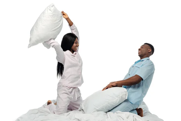 Feliz africano americano pareja teniendo almohada lucha en cama en pijama aislado en blanco - foto de stock