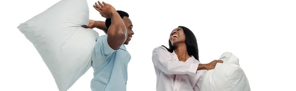 Feliz africano americano pareja teniendo almohada lucha en pijama aislado en blanco, tiro panorámico - foto de stock