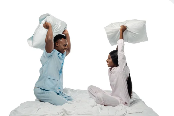 Feliz africano americano pareja teniendo almohada lucha en cama en pijama aislado en blanco - foto de stock