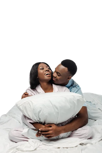 Happy african american couple hugging pillow in bed in pajamas isolated on white — Stock Photo