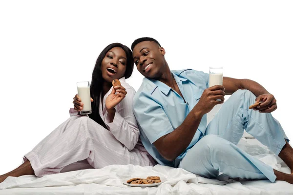 Felice coppia afroamericana mangiare biscotti con latte a letto in pigiama isolato su bianco — Foto stock