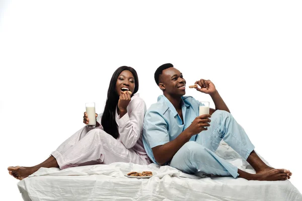 Feliz pareja afroamericana comiendo galletas con leche en la cama en pijama aislado en blanco - foto de stock