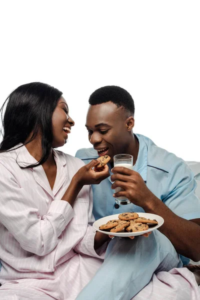 Happy african american woman feeding boyfriend with cookies in bed in pajamas isolated on white — Stock Photo