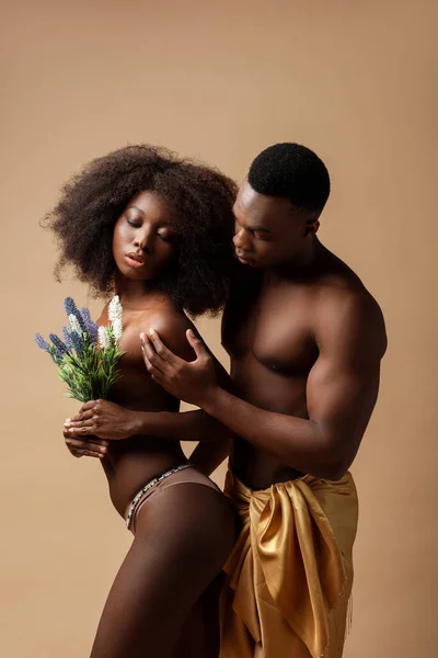 Vista lateral do casal americano africano nu sexy posando com planta isolada no bege — Fotografia de Stock