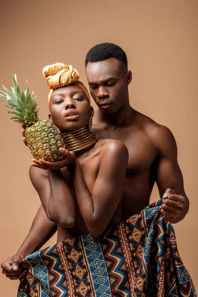 Sexy naked tribal afro woman covered in blanket posing with pineapple near man on beige — Stock Photo