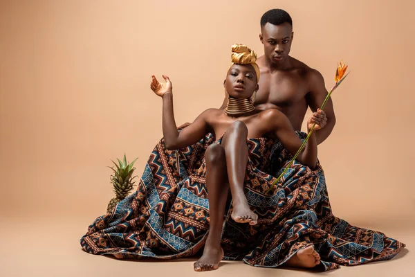 Sexy naked tribal afro woman covered in blanket posing near man and pineapple on beige — Stock Photo