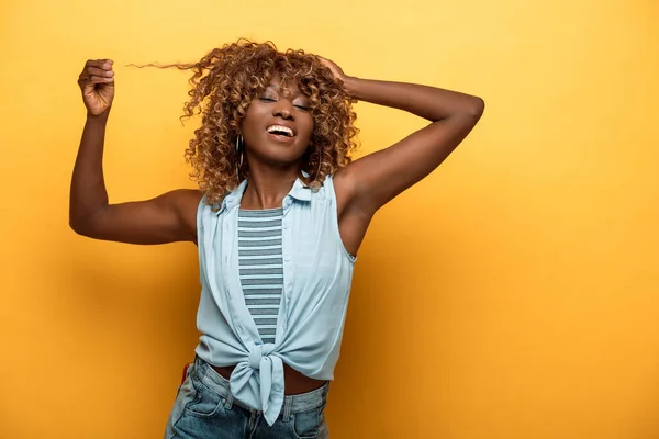 Happy african american woman touching hair on yellow background — Stock Photo