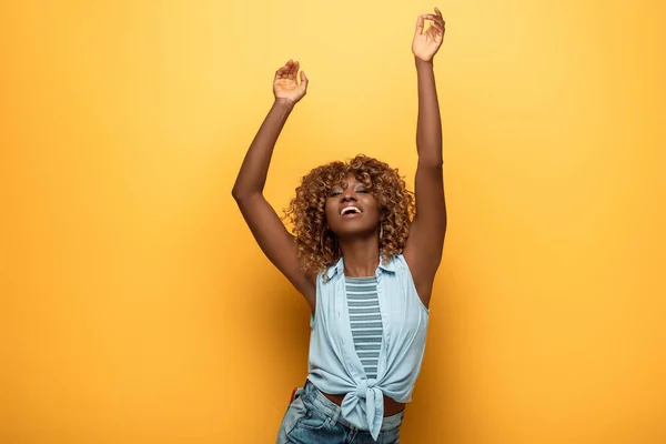 Happy african american woman dancing with hands in air on yellow background — Stock Photo