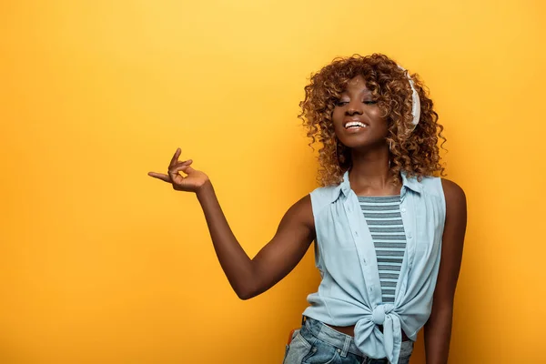 Happy african american woman listening music in headphones and pointing with finger on yellow background — Stock Photo