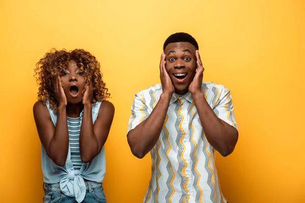 Shocked african american couple with open mouths touching faces on yellow colorful background — Stock Photo