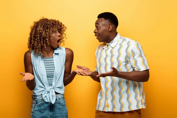 Impactada pareja afroamericana con bocas abiertas mirándose el uno al otro sobre fondo amarillo colorido - foto de stock