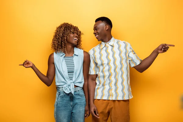 Happy african american couple pointing with fingers and looking at each other on yellow colorful background — Stock Photo