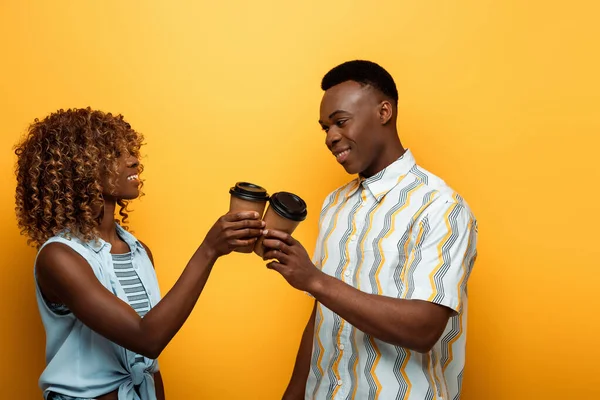 Glückliches afrikanisch-amerikanisches Paar klingelt mit Pappbechern auf gelbem, buntem Hintergrund — Stockfoto