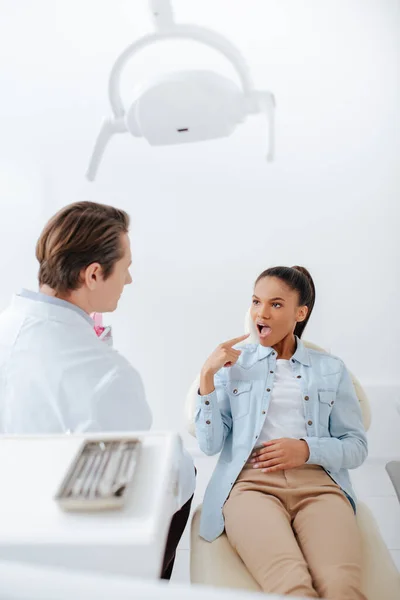 Selective focus of african american woman pointing with finger at opened mouth while looking at dentist in clinic — Stock Photo