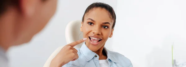 Prise de vue panoramique d'une femme afro-américaine à la bouche ouverte pointant du doigt un appareil dentaire près d'un orthodontiste en clinique — Photo de stock