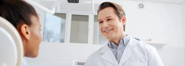 Панорамный снимок счастливого стоматолога, смотрящего на африканского американского пациента — стоковое фото