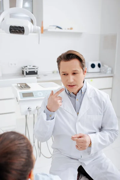 Селективный фокус стоматолога, указывающего пальцем в рот и смотрящего на африканского американского пациента — стоковое фото
