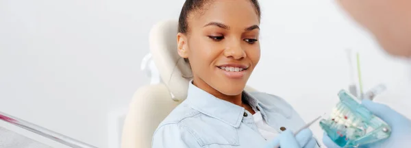 Tiro panorâmico de mulher americana africana feliz em aparelho perto dentista segurando dentes modelo — Fotografia de Stock