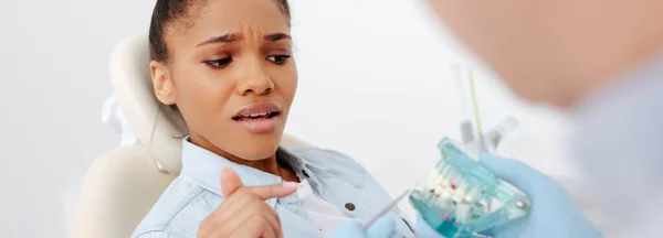 Panoramic shot of scared african american woman in braces pointing with finger at teeth model near dentist — Stock Photo