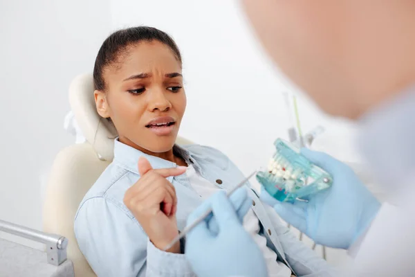 Enfoque selectivo de la mujer afroamericana asustada en frenos apuntando con el dedo al modelo de dientes cerca del dentista - foto de stock