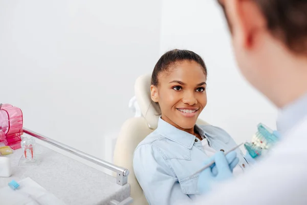 Enfoque selectivo de la mujer afro-americana alegre en frenos cerca del dentista sosteniendo los dientes modelo - foto de stock