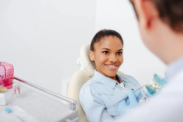 Messa a fuoco selettiva di allegra donna afro-americana in bretelle vicino al dentista con modello di denti — Foto stock